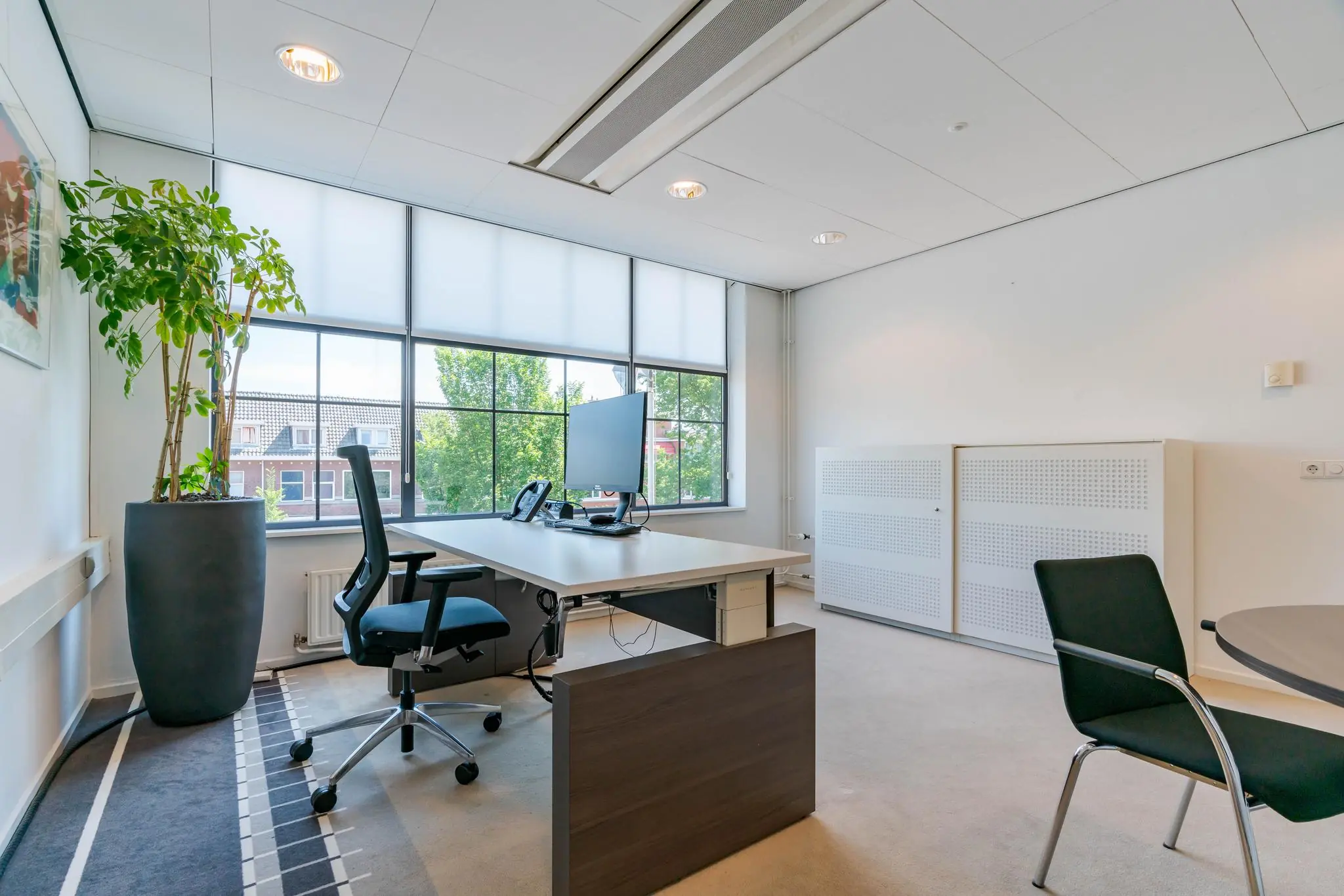 Modern en licht kantoor aan de Van Alkemadelaan met bureau, bureaustoel, vergadertafel en grote raamkozijnen.